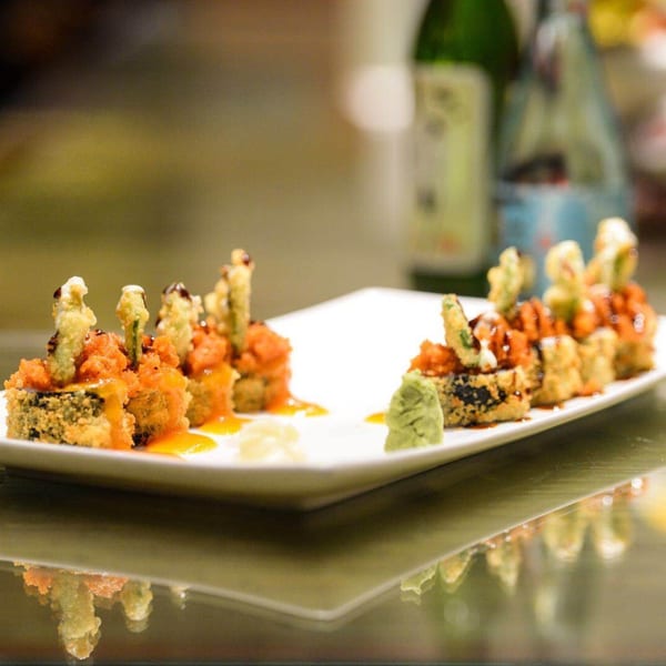 Crispy fried sushi rolls with orange topping, drizzled sauce, wasabi, and ginger on a white plate.
