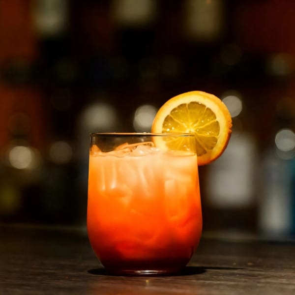 A vibrant orange cocktail with ice and an orange slice garnish sits on a dark bar.
