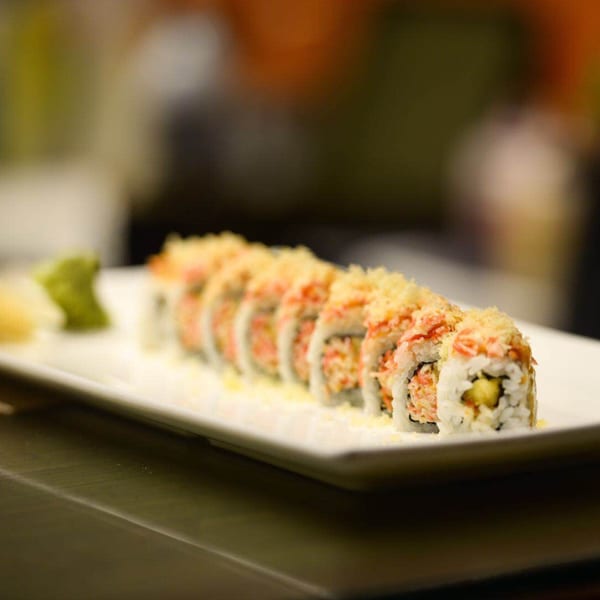 A long white plate holds a row of sushi rolls topped with crunchy tempura flakes, with wasabi and ginger.